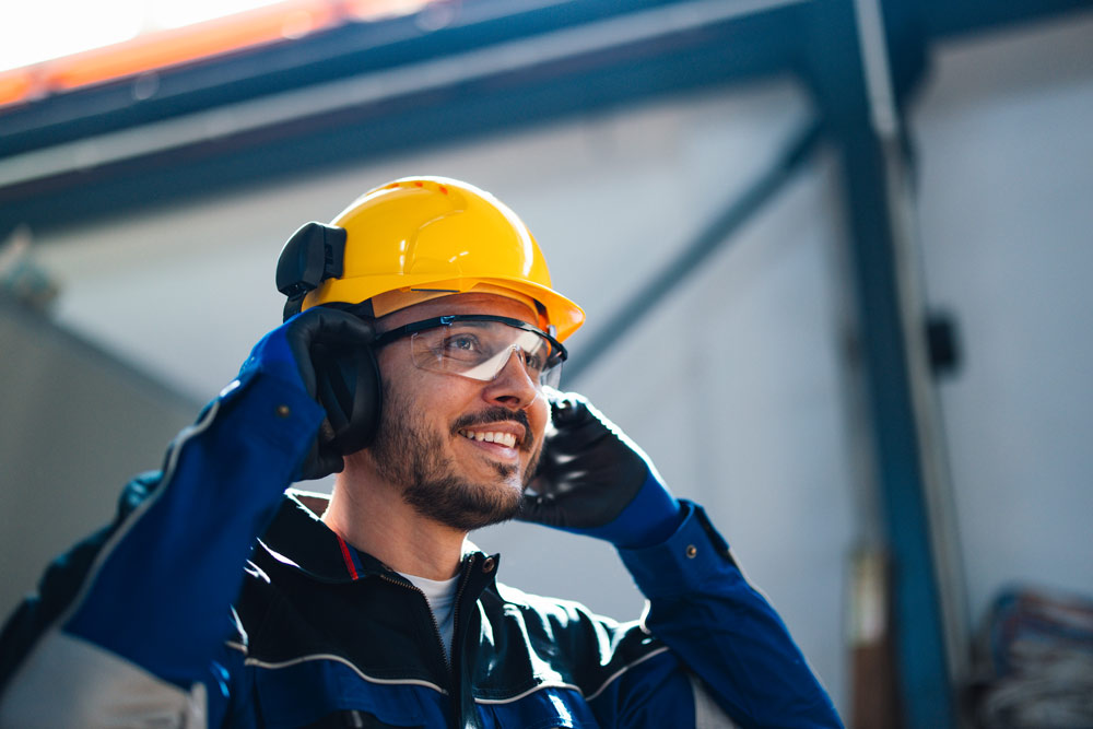 Mann mit gelbem Schutzhelm, Gehörschutz, Schutzbrille und blauen Schutzhandschuhen hebt eine Hand zum Kopf; im Hintergrund eine industrielle Stahlkonstruktion.