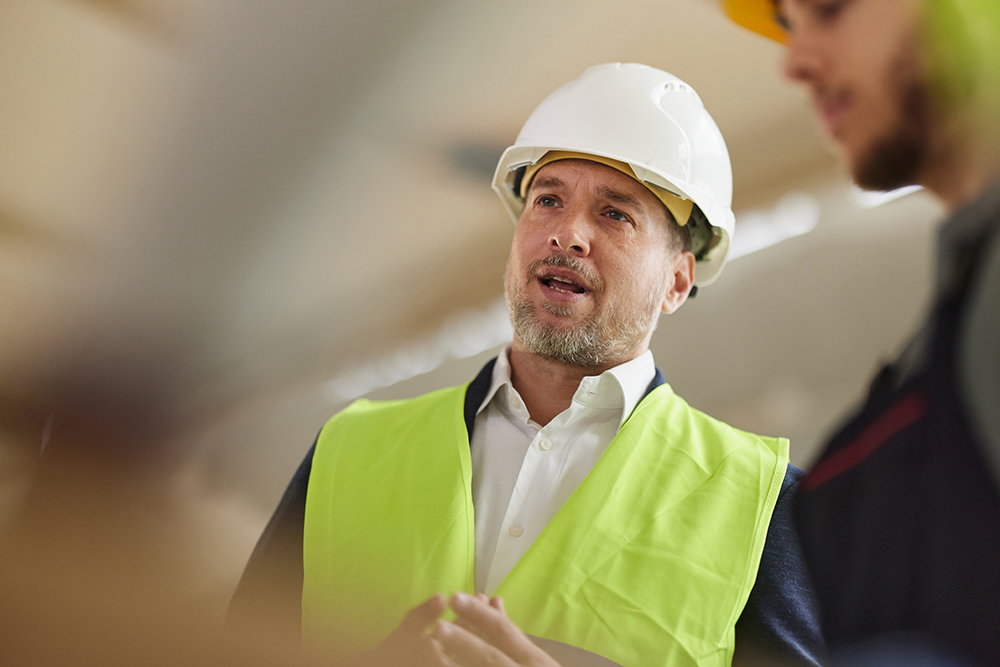 Mann mit weißem Schutzhelm und neongelber Warnweste spricht, den Blick leicht nach oben gerichtet, in einer industriellen Umgebung. Im Hintergrund rechts eine weitere Person mit gelbem Helm.