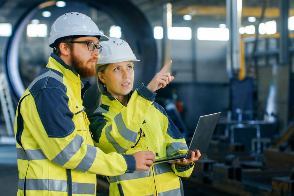 Zwei Personen in gelb-blauer Schutzkleidung und weißen Schutzhelmen stehen in einer Industriehalle. Die vordere Person hält einen aufgeklappten Laptop und zeigt mit dem Zeigefinger nach oben-rechts; die hintere Person blickt in dieselbe Richtung.