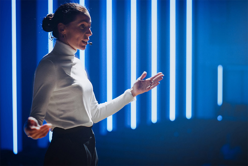 Eine Frau steht vor einem Bühnenhintergrund aus vertikalen, leuchtend weißen Lichtstreifen auf tiefblauem Grund und spricht in ein Headset-Mikrofon. Sie trägt einen weißen Rollkragenpullover und dunkle Hose, hält beide Hände in einer gestikulierenden Bewegung und blickt nach rechts.