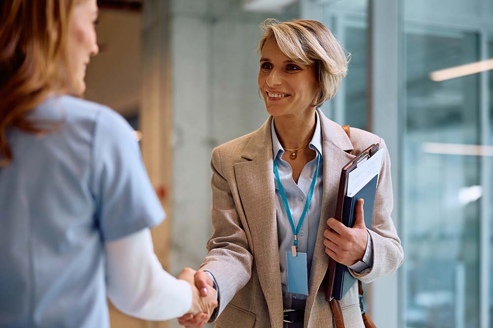 Zwei Frauen reichen sich die Hand in einem hellen Innenraum. Die vordere Person trägt einen beigen Blazer, ein türkisfarbenes Lanyard mit Ausweis und hält einen Klemmbrett-Stapel mit Dokumenten unter dem Arm.