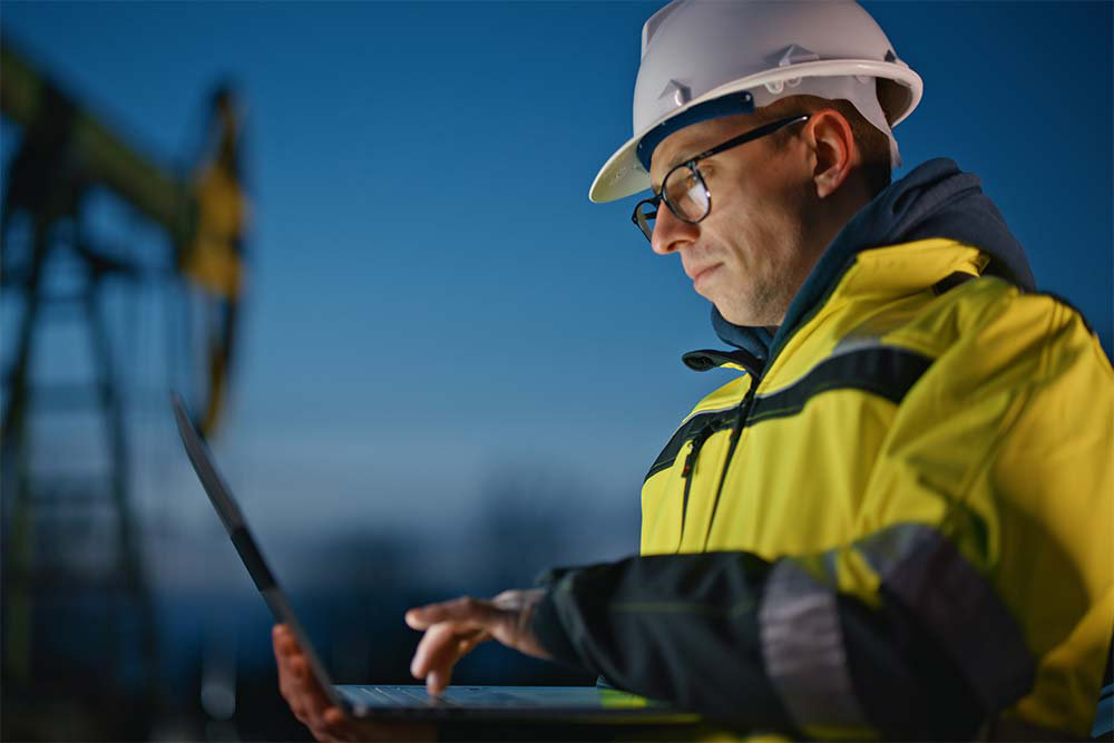 Mann mit weißem Schutzhelm und gelb-schwarzer Warnschutzjacke blickt auf einen aufgeklappten Laptop, im unscharfen Hintergrund eine Förderanlage (Pumpjack) unter blauem Abendhimmel.