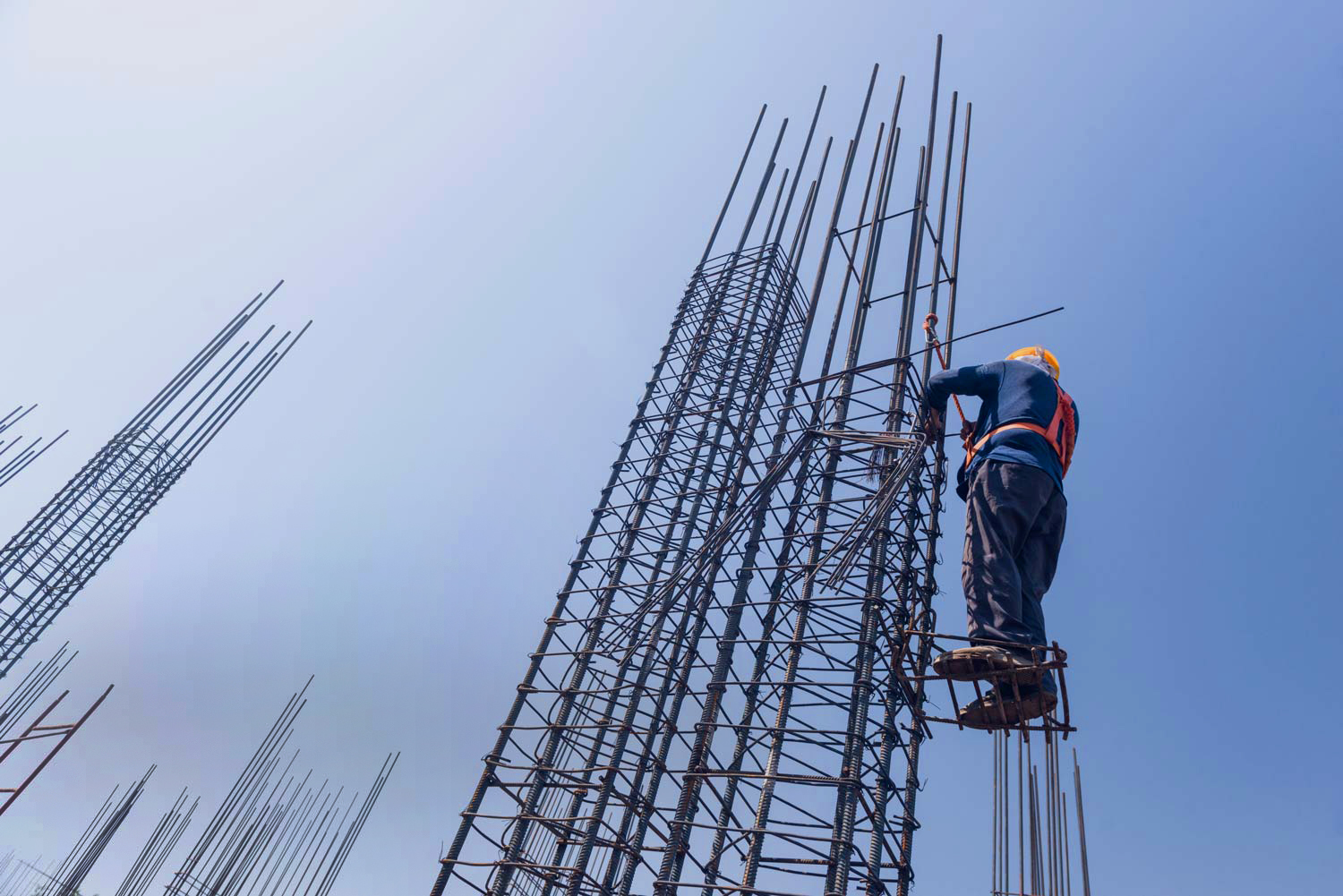 Bauarbeiter in orangefarbener Schutzweste und gelbem Helm arbeitet hoch oben an einem dichten Geflecht aus senkrecht aufragenden Stahlbewehrungsstäben auf einer Baustelle.