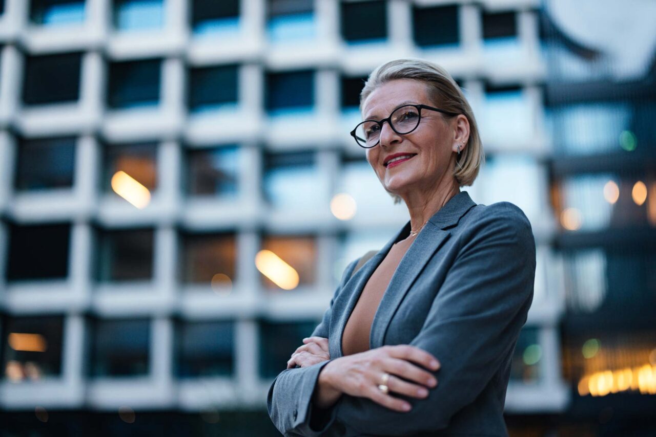 Frau mittleren bis höheren Alters mit kurzen blonden Haaren und schwarzer Rundbrille, in grauem Blazer, Arme verschränkt, leicht zur Seite blickend – vor einem unscharf bokeh-gerenderten Glasfassaden-Gebäude in Abenddämmerung.